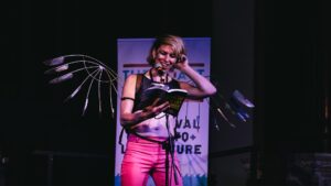 A performer at the coast is queer. Reading on stage wearing a pair of wire wings.