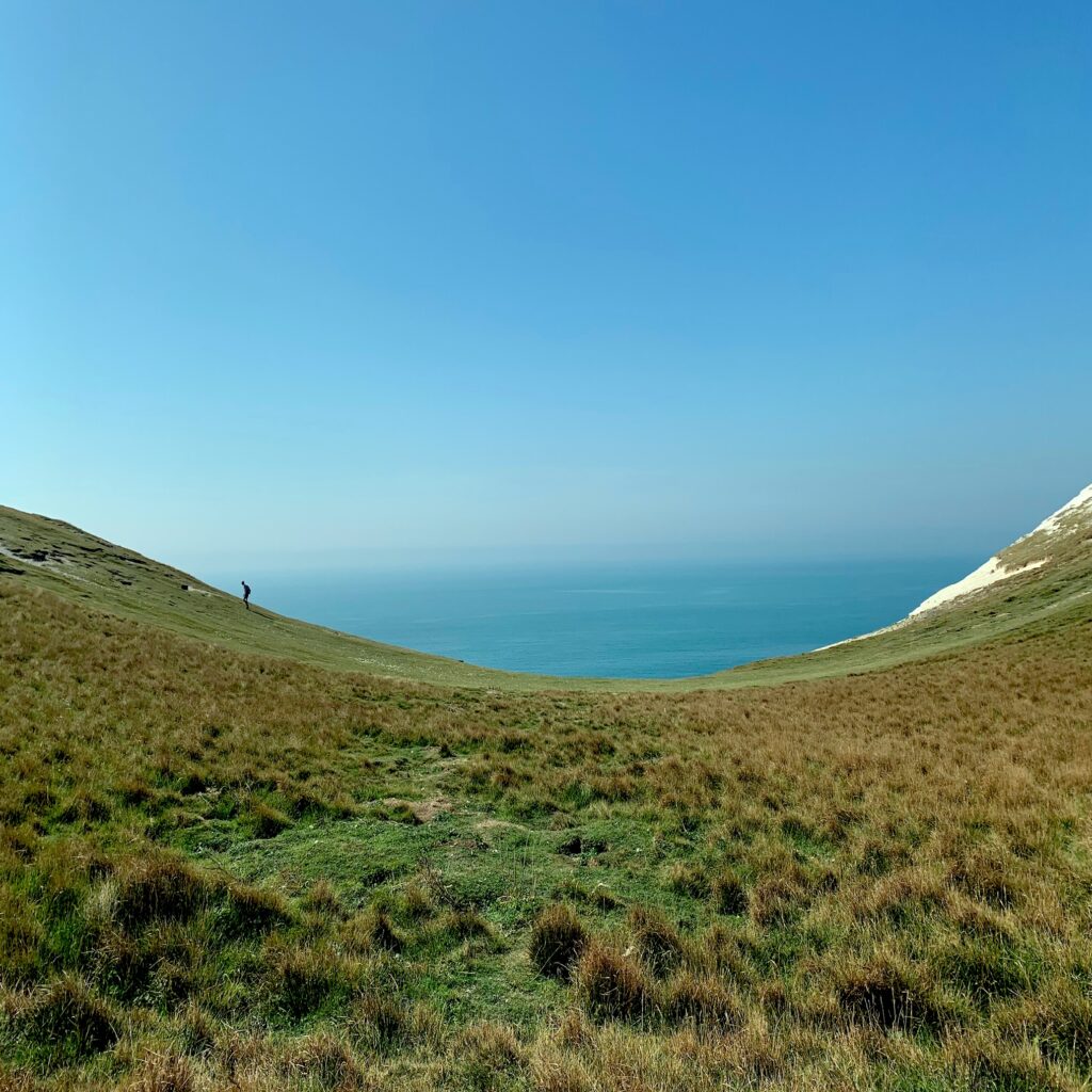 A photograph of Birling Gap, a dramatic and scooped-out dip between two tall cliffs. The sky is clear blue and the ground is covered in rich green grasses.